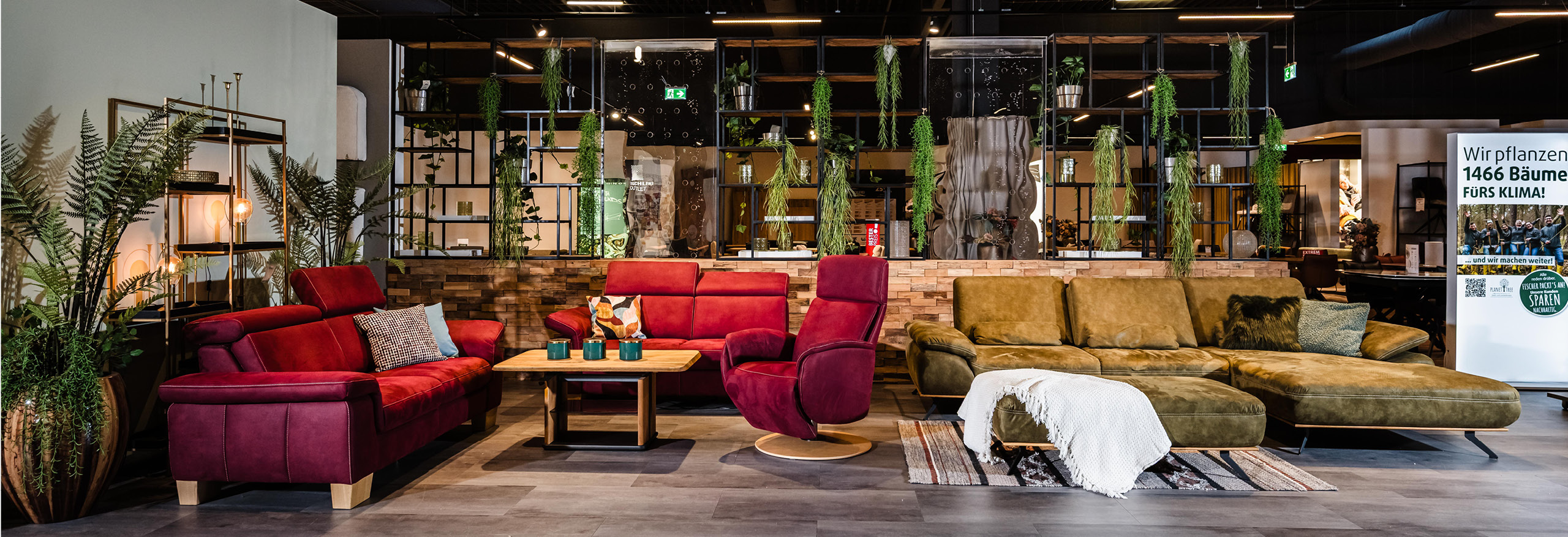 Wohnlandschaft-Ausstellung bei Polstermöbel Fischer Bruchsal mit rotem Sofa-Set, grünem Designersofa und vielen Hängepflanzen vor rustikaler Steinwand.