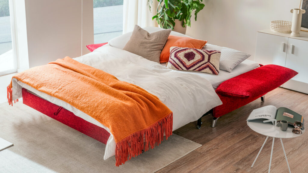 Rotes Schlafsofa im ausgezogenen Zustand mit kuscheliger Bettdecke – funktionales Schlafsofa mit Gästebettfunktion für kleine Räume.