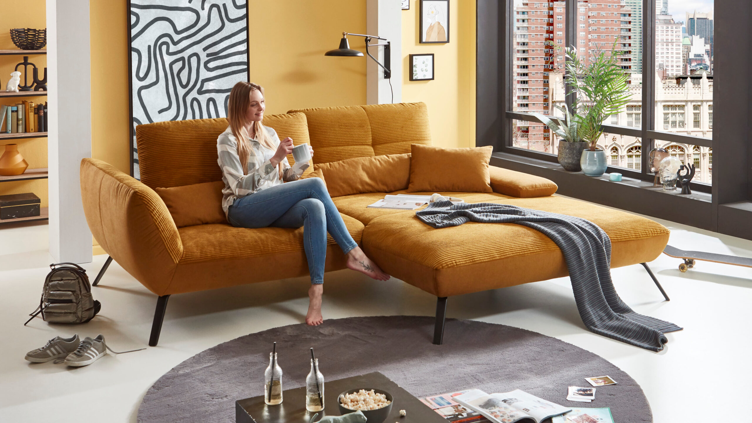 Trendiges Relaxsofa in goldgelbem Cordstoff mit lockerer Einrichtung und dekorativer Wandgestaltung – perfekter Rückzugsort für moderne Stadtwohnungen.