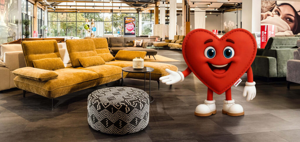 Herz-Maskottchen von Polstermöbel Fischer im modernen Möbelhaus vor gelbem Sofa – einladende Wohnlandschaften und Sofas im Showroom entdecken